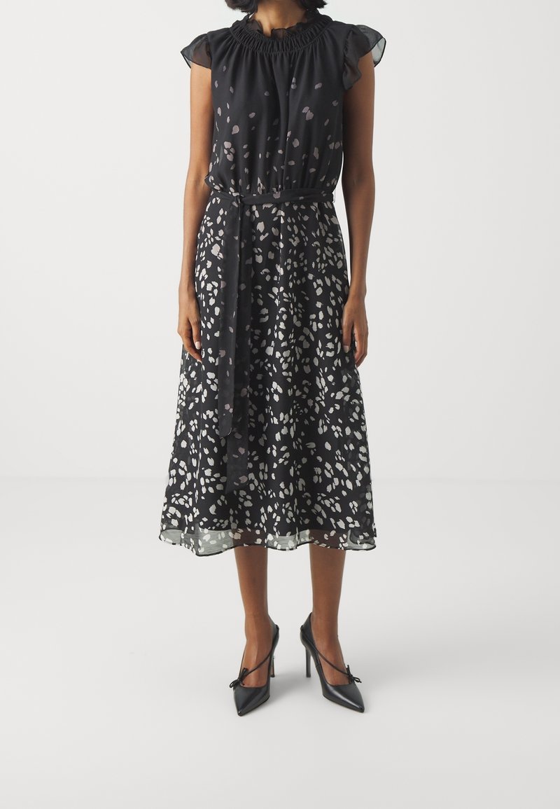 Femme portant une robe midi noire avec des taches abstraites blanches, un col volanté, des manches courtes, une taille nouée et des talons hauts noirs pointus.
