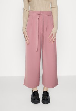 Frau trägt hochgeschnittene, weit geschnittene pinke Hose mit gebundenem Gürtel, beigen, gerippten Langarmshirt und schwarze Loafers, steht vor weißem Hintergrund.