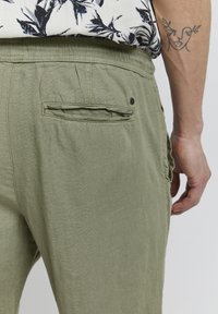 Pantalons légers verts, coupe décontractée, en tissu texturé, avec taille élastique et poche arrière. Présente des coutures décoratives et un accent de bouton.