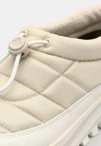 Zapatilla beige acolchada con suela blanca lisa de goma. Cuenta con un cierre de botón y parte superior textil suave, que ofrece un aspecto acolchado.
