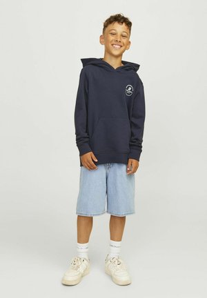 Sötétkék kapucnis pulóver, elől zsebbel, világoskék farmernadrággal és fehér sneaker-rel viselve, a mellrészén logóval.