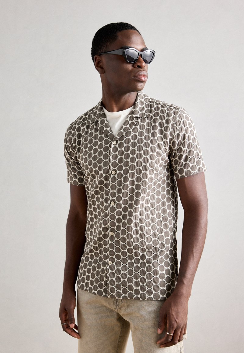 Camisa de mangas curtas, com colarinho, com um padrão geométrico em tons de castanho e creme. Tecido texturizado, leve. Acompanhada de óculos de sol pretos.