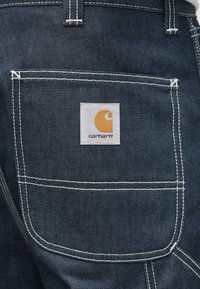 Σκούρο τζιν παντελόνι εργασίας με πίσω τσέπη που φέρει μια λευκή τετράγωνη ετικέτα με πορτοκαλί λογότυπο και λευκές λεπτομέρειες ραφής.