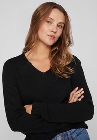 Svart v-ringad tröja med långa ärmar, tillverkad av ett mjukt stickat material. Har ribbade muddar och nederkant, bärs över denimjeans.
