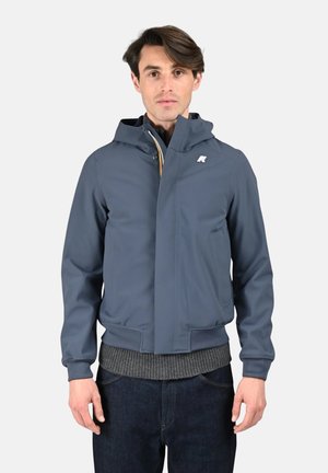 Homme portant une veste à capuche bleue par-dessus un pull gris et un jean foncé, debout face à l'avant sur un fond uni.