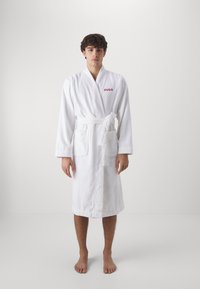 Weißer Bademantel aus Frottee mit Schalkragen, Bindegürtel und zwei frontalen Taschen; trägt das rote "HUGO"-Logo auf der Brust.