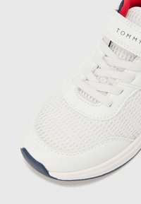 Balti sportiniai bateliai iš tinklelio su tekstūruotu viršumi, apvalia nosimi, pilkos gumos padu ir raudonu vidiniu pamušalu su balta dirželiu, ant kurio parašyta „TOMMY“.