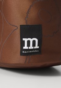 Brown fabric bag featuring a floral pattern in darker brown. Black label with white text: "m marimekko." Smooth texture, rounded shape.