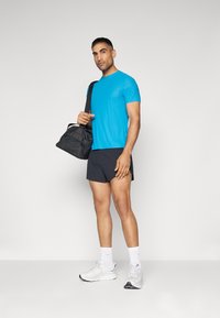 Conjunto atlético masculino com uma t-shirt de mangas curtas azul brilhante, calções pretos e sapatilhas brancas, a segurar uma mala de desporto preta.