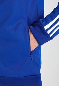 Blauwe sportjack met een ritszak aan de linkerkant, geribbelde manchetten en drie witte strepen langs de mouw voor detail.