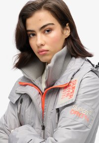 Lys grå jakke med høj krave, orange lynlåse og logo-patches. Har lynlåsede lommer og detaljeret ribbet krave.