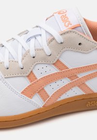 Vitt sportskor med ljusorange accenter, mockaöverlägg och platta vita snören. Har perforeringar för andningsförmåga och en gummisula.