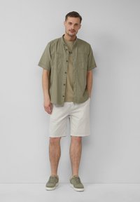 Olijfgroen overhemd met korte mouwen over een beige t-shirt, gecombineerd met lichte gestreepte shorts en olijfgroene sneakers. Eenvoudige, casual outfit.