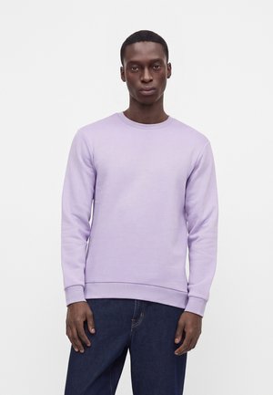 Mann trägt einen schlichten hellvioletten Sweatshirt mit Rundhalsausschnitt und dunkelblaue Jeans, steht vor weißem Hintergrund.