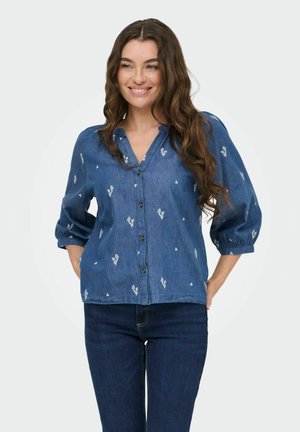 Femme aux longs cheveux ondulés souriante, portant un chemisier bleu à boutons avec des imprimés floraux blancs et un jean foncé, les mains glissées derrière.