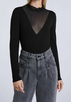 Femme portant un haut noir à manches longues avec un col en V transparent et un jean gris plissé à taille haute avec passants pour ceinture.