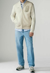Felpa zip leggera color beige chiaro con una toppa a forma di lettera "B" e testo ricamato, indossata sopra una camicia a righe blu chiaro e jeans blu.