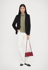 Donna con lunghi capelli neri, vestita con un blazer nero, una camicia a righe verde oliva, pantaloni bianchi e ballerine nere, che tiene una borsa rossa per la maniglia.