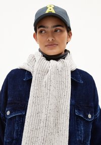 Hellgrauer, gerippter Strickschal mit strukturiertem Muster, getragen über einer marineblauen Cordjacke. Die Mütze hat ein gelbes Logo auf der Vorderseite.