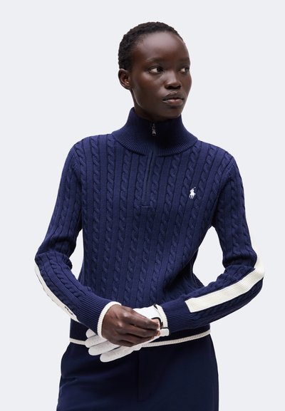 Femme aux cheveux courts portant un pull en maille torsadée bleu marine avec des rayures blanches aux manches, tenant des gants blancs devant un fond uni.