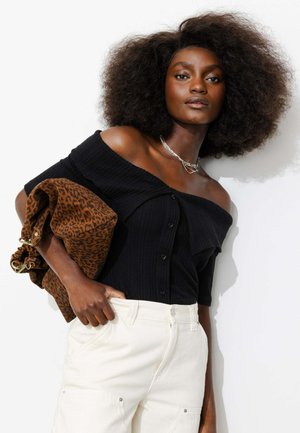 Femme aux cheveux afro naturels, portant un top noir épaules dénudées et un pantalon blanc, tenant un grand sac marron à imprimé léopard.