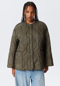 Person wearing olive green quilted jacket with front buttons and two pockets, paired with light blue jeans and braided hair.