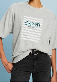 Ljusgrå oversize T-shirt med randigt mönster och texten "ESPRIT NEW YORK". I kombination med svarta jeans och ett bälte med kedjedetaljer.