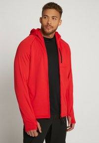 Giacca a zip rossa con cappuccio e maniche lunghe, dotata di una tasca con zip sul davanti e accenti neri. Realizzata in tessuto liscio e elasticizzato.