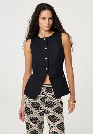 Vrouw met lang krullend haar, gekleed in een mouwloos zwart vest met knopen en een broek met een patroon van grote zwarte vissen.