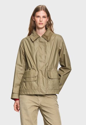 Jeune femme portant une veste beige avec un col en velours côtelé et un pantalon beige assorti sur un fond uni clair.
