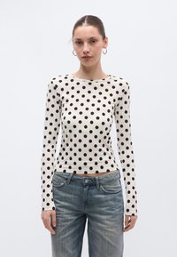 Pitkähihainen pusero kermanvärisenä, jossa suuret mustat pilkut, pehmeä materiaali, pyöreä pääntie, yhdistetty vaaleisiin farkkuihin.