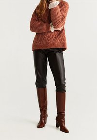 Pull en maille couleur rouille avec un motif texturé, pantalon en cuir synthétique noir et bottes marron à talons montantes jusqu'aux genoux.