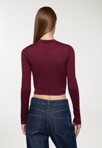 Top a maniche lunghe color bordeaux con un design aderente, abbinato a jeans in denim blu a vita alta. Tessuto morbido con una leggera lucentezza.