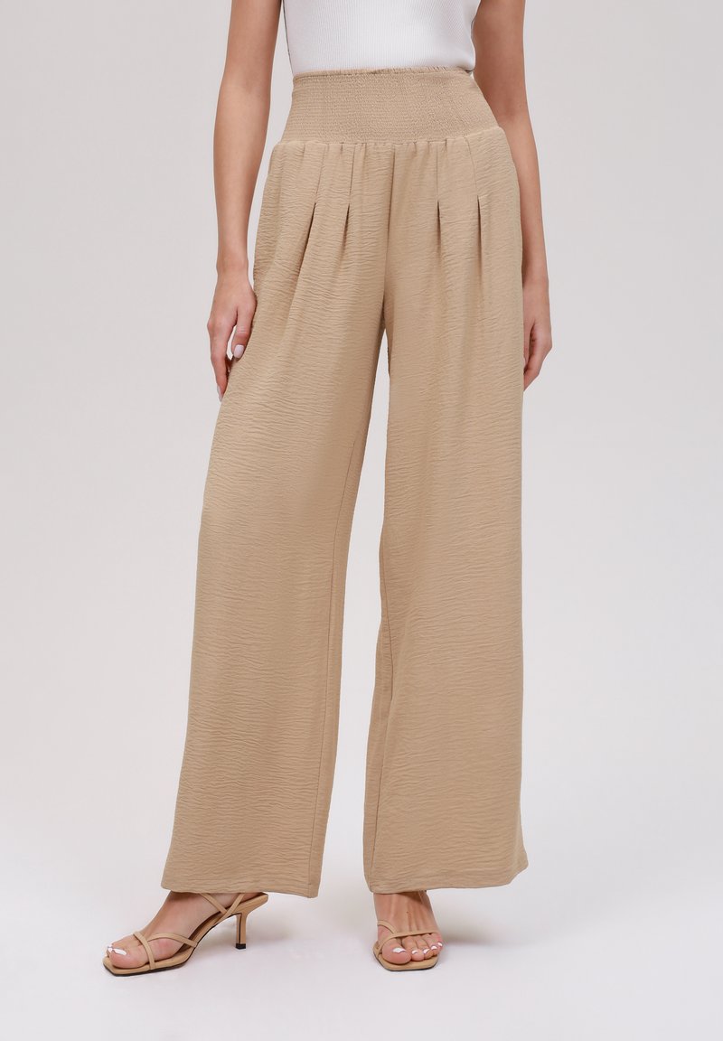 Beżowe spodnie z szerokimi nogawkami o teksturowanym wykończeniu, z marszczonym pasem i plisami z przodu. Połączone z beżowymi sandałkami na paski.