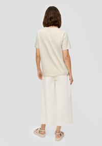 Hellbeige Baumwoll-T-Shirt mit kurzen Ärmeln, kombiniert mit weiten weißen Hosen. Die Schuhe haben beige Riemen und eine Plateausohle.