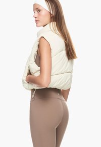 Pufferväst i ljusbeige med kviltad design. Har cropped längd och sidodragband. Modellen bär bruna leggings. Minimalistisk stil.
