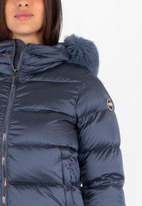 Giubbotto puffer blu navy con cappuccio rifinito in pelliccia, presenta pannelli trapuntati orizzontali e chiusura con zip. Decorato con un logo sulla spalla.