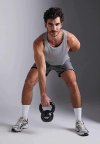 Vyras šviesiai pilku marškinėliu be rankovių ir šortais atlieka kettlebell pakėlimą. Kettlebell yra juodas su svorio žyme, batai yra sidabriniai ir balti.