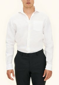 Camicia di lino bianca con tasca sul petto, polsini bottonati e bottoni chiari, abbinata a pantaloni tailored scuri. Tessuto di consistenza liscia.