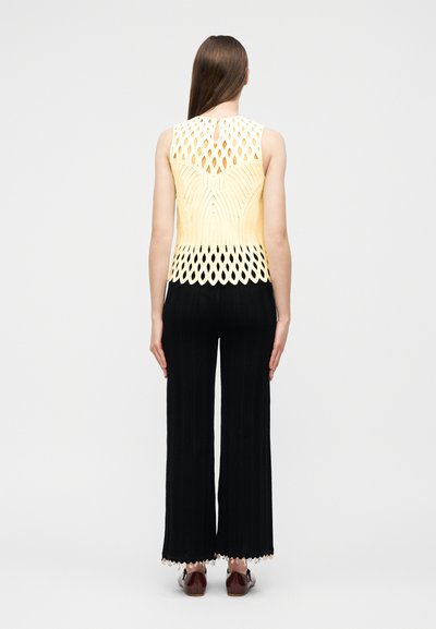 Femme portant un haut jaune sans manches avec des motifs découpés et un pantalon noir à jambes larges, debout de dos sur un fond clair uni.