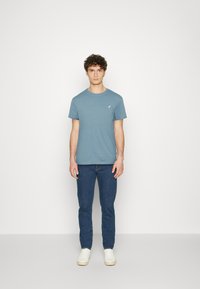 Blue cotton t-shirt with a crew neck and short sleeves, featuring a small white logo on the chest. Paired with blue jeans and white sneakers.