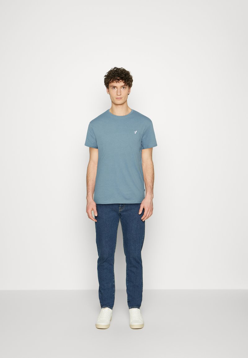Blue cotton t-shirt with a crew neck and short sleeves, featuring a small white logo on the chest. Paired with blue jeans and white sneakers.