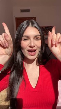 Pikkade tumejate juustega naine punases särgis, naeratades ja tõstes mõlemad nimetissõrmed ülespoole heleduse ja loomuliku sisevalgustuse keskel.