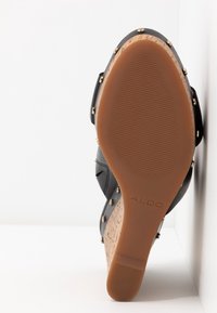 Sandale noire avec un talon en liège, une semelle en caoutchouc et des clous dorés sur le dessus. Le design intègre un motif texturé.