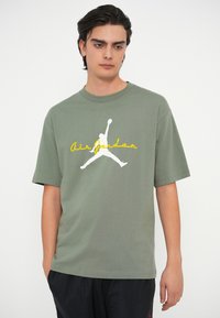 Ελαιόλαδο πράσινη μπλούζα φτιαγμένη από βαμβάκι, με λευκό λογότυπο σιλουέτας Jordan και κίτρινο κείμενο "Air Jordan" στο στήθος. Κοντό μανίκι.