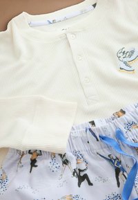 Kreminės spalvos raukšlėta ilgomis rankovėmis palaidinė, derinama su šviesiai mėlynomis raštuotomis kelnėmis, kuriose pavaizduoti čiuožėjai, su elastinga juosmeniu ir mėlyna raišteliu.