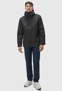 Zwarte waterdichte jas met een hoge kraag, rits aan de voorkant en verstelbare capuchon. Draagt over blauwe jeans en witte sneakers.