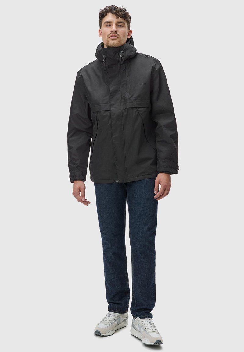 Zwarte waterdichte jas met een hoge kraag, rits aan de voorkant en verstelbare capuchon. Draagt over blauwe jeans en witte sneakers.