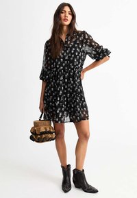 Black floral dress with sheer fabric, puff sleeves, and a tiered skirt. Black studded ankle boots and a textured woven handbag.