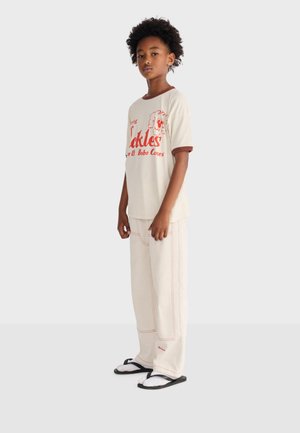 Barn med krøllet hår iført en beige T-shirt med rød tekst og illustrationer, beige bukser med rød syning samt sorte sandaler med hvide sokker.